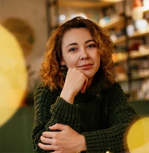 A woman in a green sweater resting her chin on her hand, smiling softly in a warmly lit café